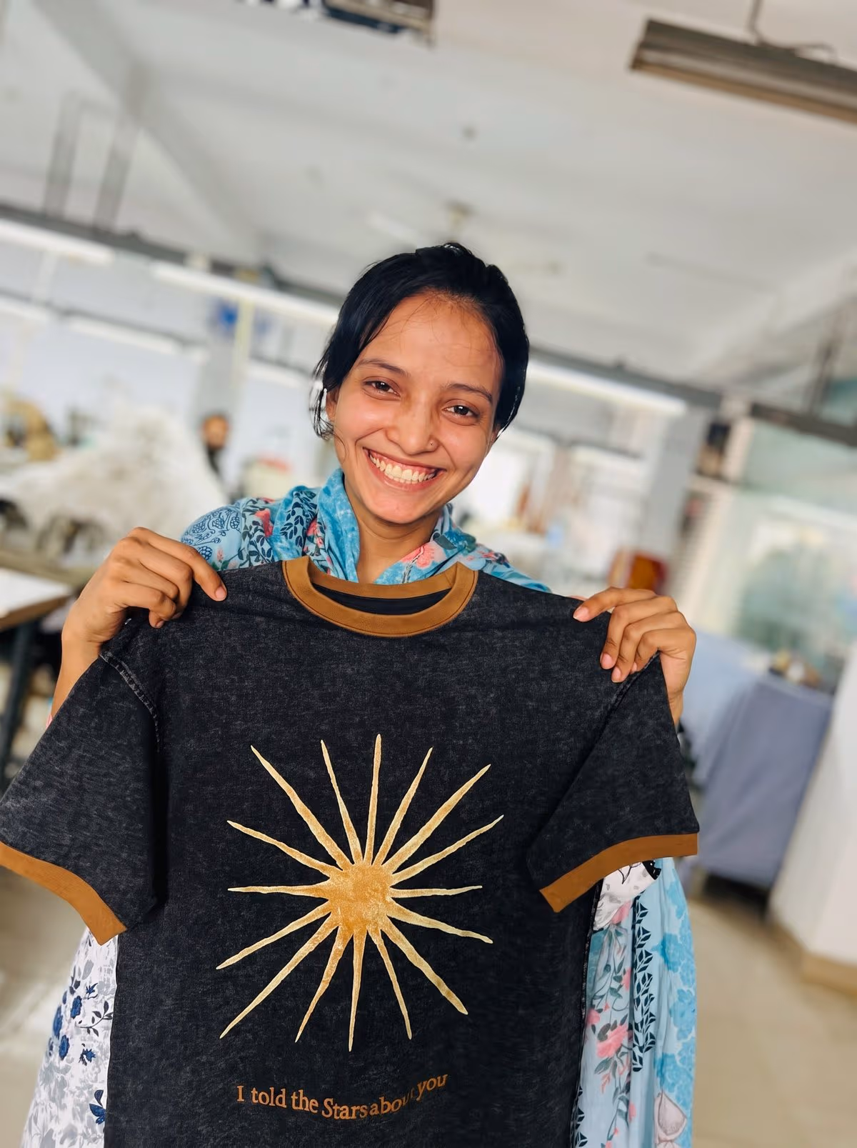 A Collective Studio team member smiling and holding up a finished charcoal T-shirt with a gold sunburst print and the text "I told the Stars about you."